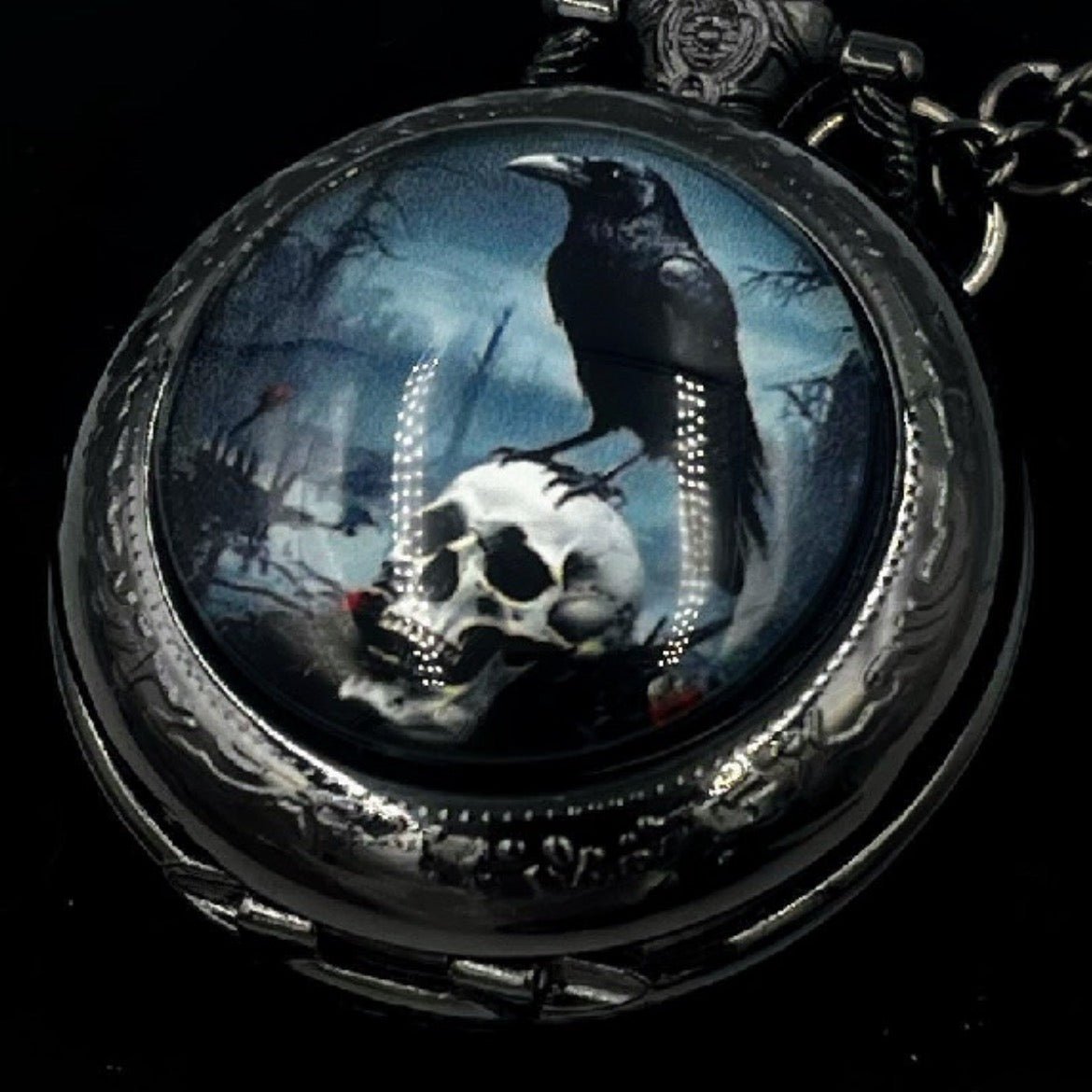 A gothic-style pocket watch pendant featuring a detailed illustration of a black raven perched on a white skull against a dark blue and black background, set in a silver-toned metal case with a matching chain.