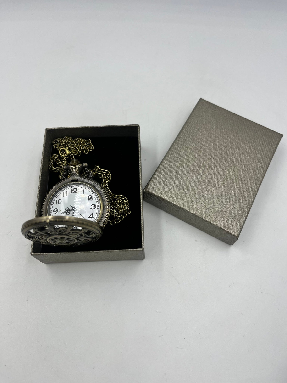 Open bronze-tone pocket watch showing a white numbered dial, with chain and box lid alongside, Steampunkgearsa.