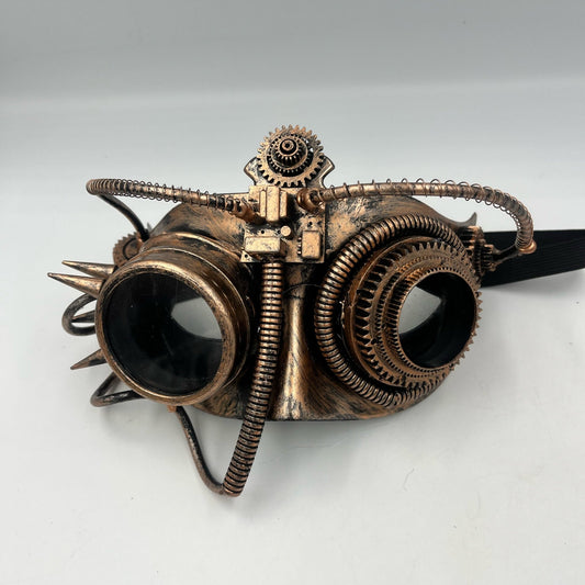 Steampunk-style metal goggles with two large dark lenses, exposed gears, springs, spikes, and a black strap, photographed on a plain light background.